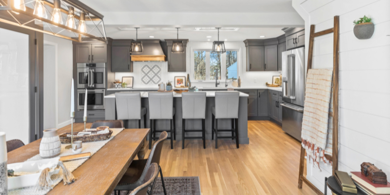 A spacious kitchen and dining area with gray cabinetry, a central island with modern bar chairs, pendant lighting, and stainless-steel appliances. A wooden dining table with benches and chairs sits adjacent to the kitchen.