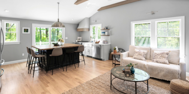 An airy open space combining a kitchen with gray cabinetry and stainless steel appliances, a dining area with a dark round table and chairs, and a living area with a comfortable couch and glass coffee table, all over light hardwood floors