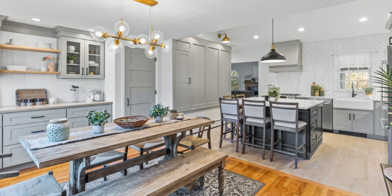 A bright kitchen renovation with gray cabinetry, white countertops, a central island with bar seating, unique globe chandeliers, and a rustic wooden dining table paired with a bench and chairs on a patterned area rug