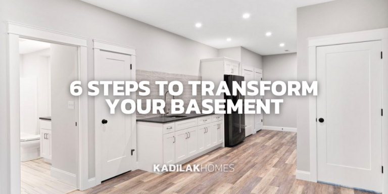 A sleek basement conversion featuring a kitchenette with white cabinetry, dark countertops, and stainless steel appliances. The space is well-lit with recessed ceiling lights and has the same hardwood flooring and white walls as the previous image. A closed white door suggests additional rooms. Text overlay in bold reads '6 STEPS TO TRANSFORM YOUR BASEMENT', positioned above the 'KADILAK HOMES' logo in the lower left corner.