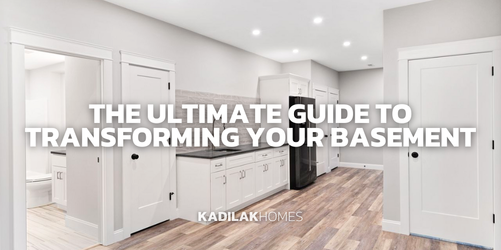 A sleek basement conversion featuring a kitchenette with white cabinetry, dark countertops, and stainless steel appliances. The space is well-lit with recessed ceiling lights and has the same hardwood flooring and white walls as the previous image. A closed white door suggests additional rooms. Text overlay in bold reads 'THE ULTIMATE GUIDE TO TRANSFORMING YOUR BASEMENT', positioned above the 'KADILAK HOMES' logo in the lower left corner.
