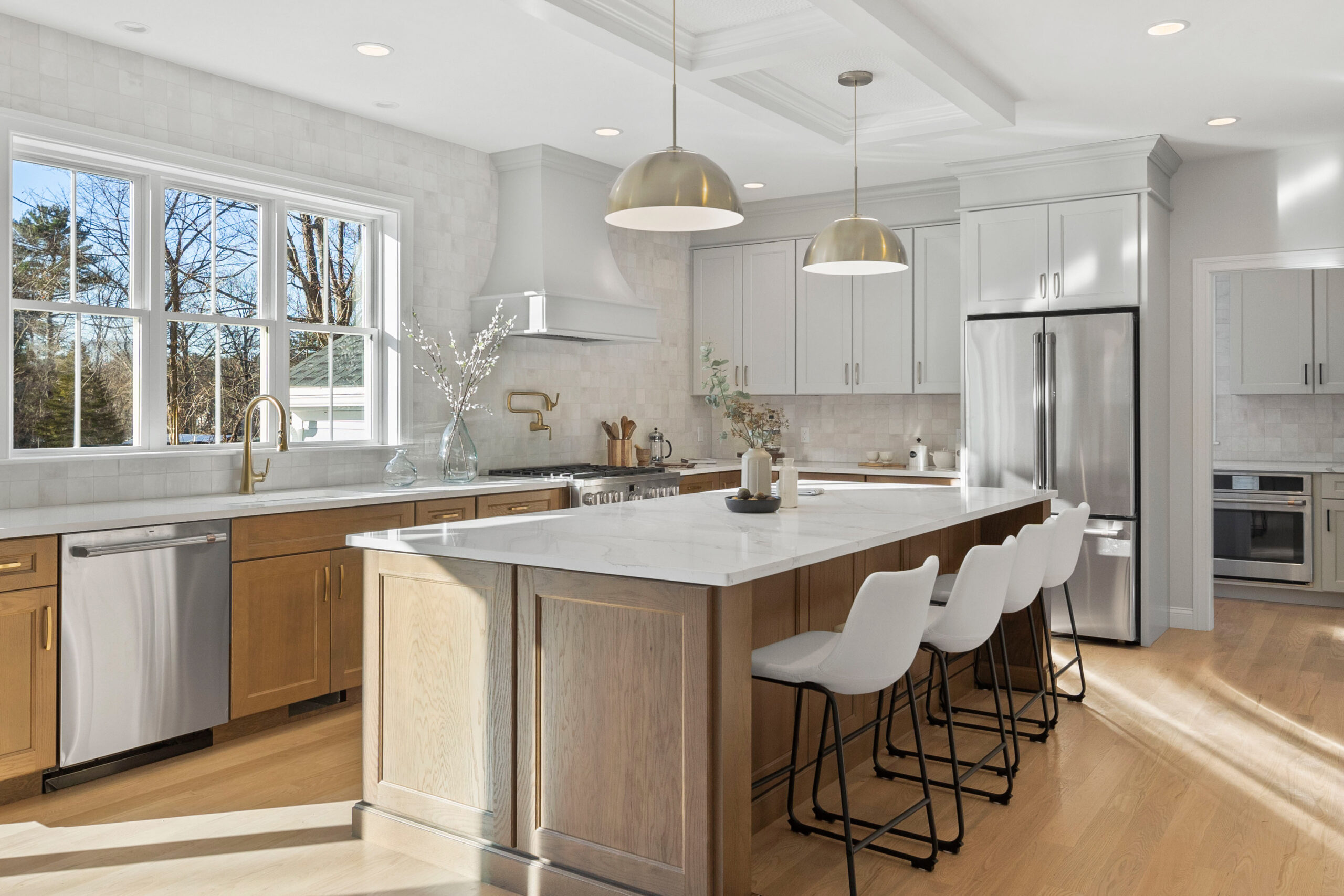 A spacious and modern kitchen with abundant natural light streaming through large windows that offer a view of bare trees and a clear sky. The kitchen features sleek white cabinetry and stainless steel appliances, including a refrigerator and built-in microwave. The centerpiece is a large kitchen island with light wood base cabinets and a white marble countertop, complemented by four white modern bar stools with black metal legs. Above the island, two large dome-shaped gold pendant lights add a touch of elegance. The backsplash is made of subway tiles, and gold fixtures and cabinet pulls match the lighting. The room has a coffered ceiling with recessed lighting and the floor is laid with light hardwood, reflecting the sunlight beautifully.