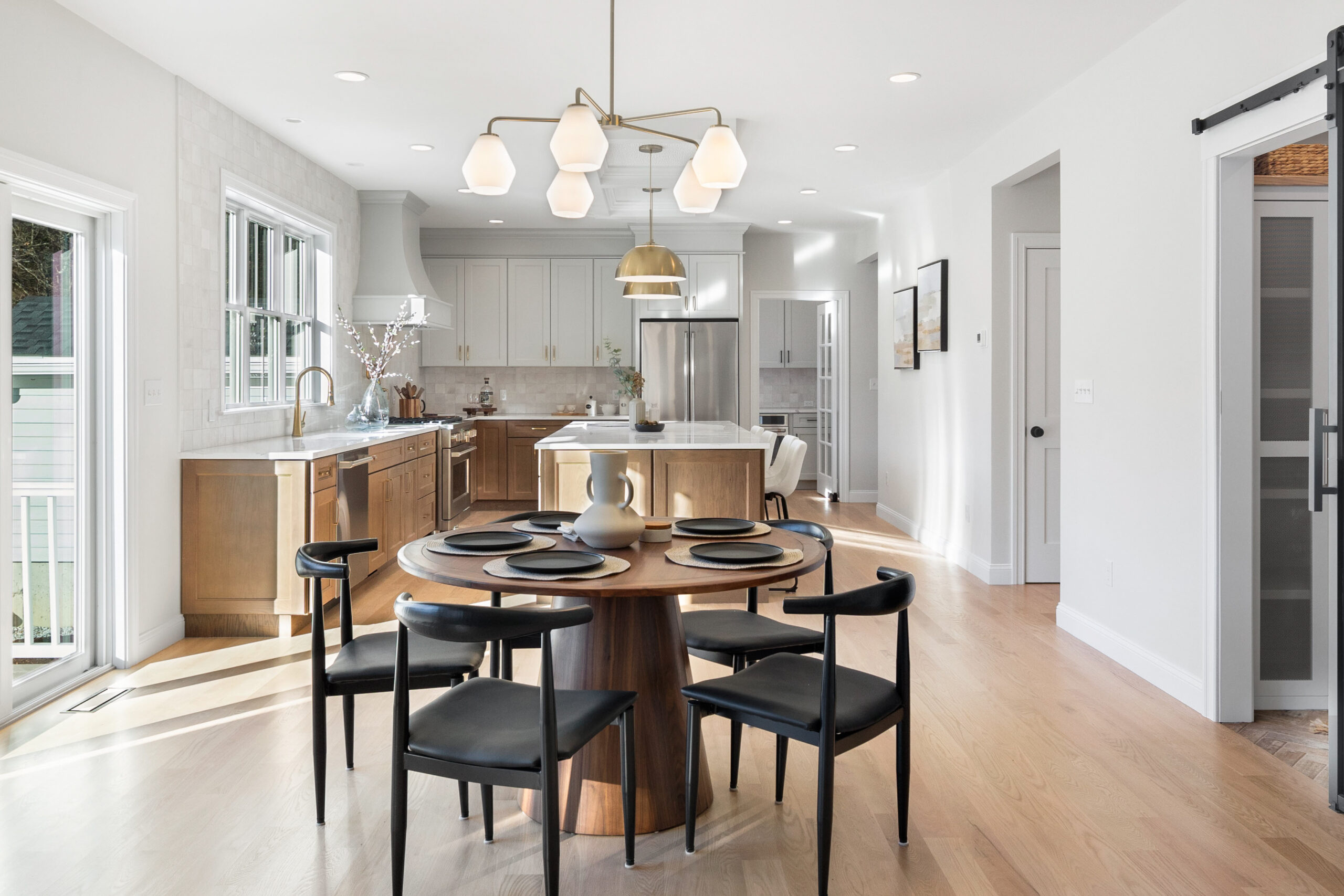 A well-lit open-plan space combining a dining area and kitchen. The dining area features a round, dark wood table with contemporary black chairs, set under a chic chandelier with multiple milky glass shades. In the kitchen area, white cabinetry with sleek hardware matches the marble countertops, and a central island provides additional seating with high stools. Two gold pendant lights add a touch of elegance over the island. Natural light floods the room through large windows and a sliding glass door, enhancing the warm tones of the light wood flooring. The kitchen is equipped with stainless steel appliances, and a black sliding barn door adds a modern farmhouse feel to the space.