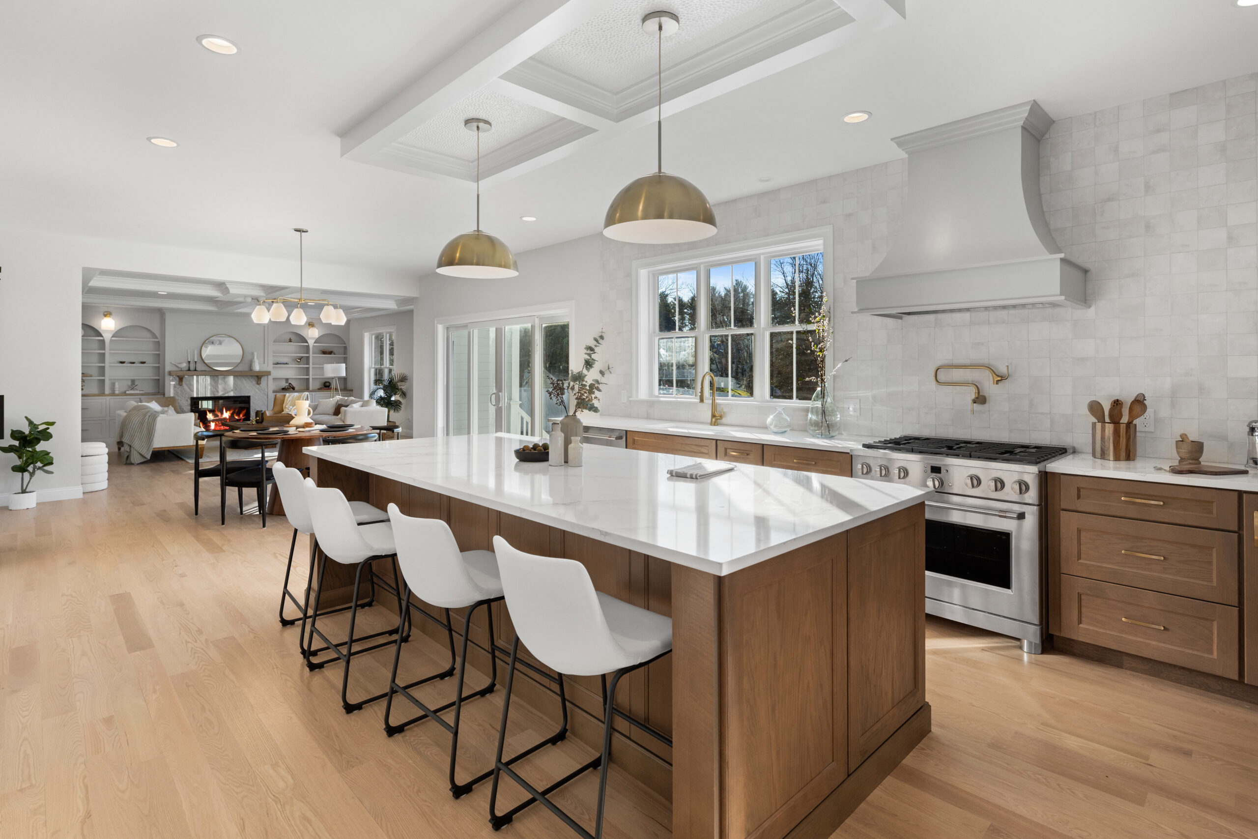 An expansive open-concept kitchen with a large center island featuring a white countertop and wooden base, providing seating for several people on sleek white stools with black legs. Gold pendant lights hang above the island, adding a touch of luxury to the space. The kitchen boasts a professional-grade stainless steel stove and oven, with a white hood and a herringbone tile backsplash. Gold fixtures and hardware complement the warm wooden tones of the cabinets. The view through the window shows a bright, clear day. Beyond the kitchen, the living area is visible, with a cozy fireplace and a relaxing seating arrangement. The entire space is unified with light hardwood flooring and a white coffered ceiling with recessed lighting.