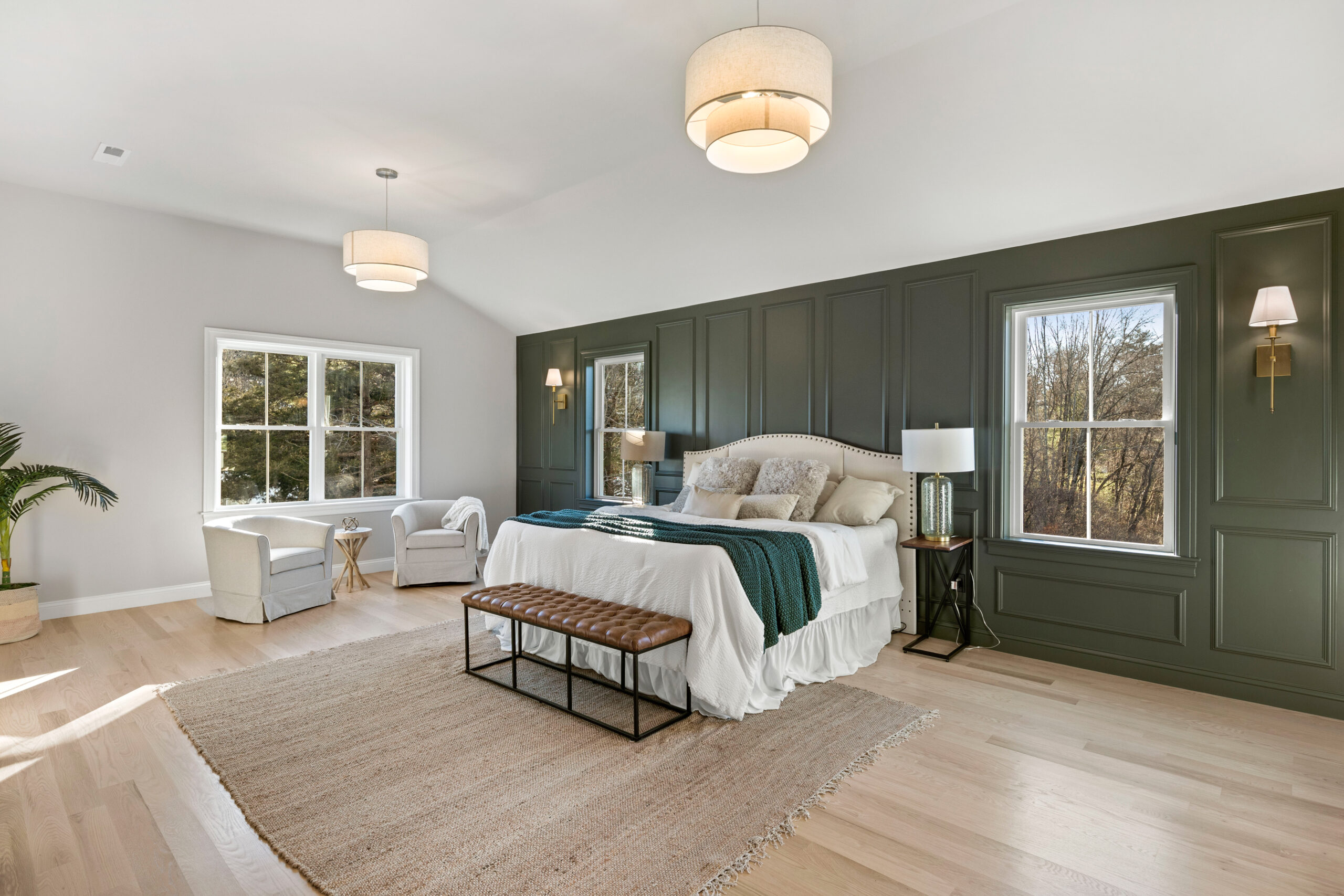 A spacious and serene bedroom with a wall of dark green wainscoting providing a dramatic backdrop to an elegant white bed adorned with plush pillows and a green throw. Two large windows allow natural light to stream in, highlighting the light hardwood floors and a large area rug. A tufted leather bench sits at the foot of the bed, and a cozy reading nook is created with two armchairs and a small side table near the window. The room features two stylish drum pendant lights, and on the far side, a thriving potted plant adds a touch of nature to the interior. This bedroom exudes a relaxed, luxurious atmosphere. Ashwood Moss 1484