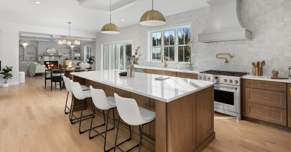 A modern kitchen featuring natural wood cabinets, white quartz countertops, and gold accents. The open floor plan connects the kitchen with a cozy living space, showcasing built-in shelving and a fireplace
