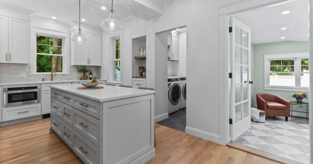 A modern kitchen featuring natural wood cabinets, white quartz countertops, and gold accents. The open floor plan connects the kitchen with a cozy living space, showcasing built-in shelving and a fireplace.