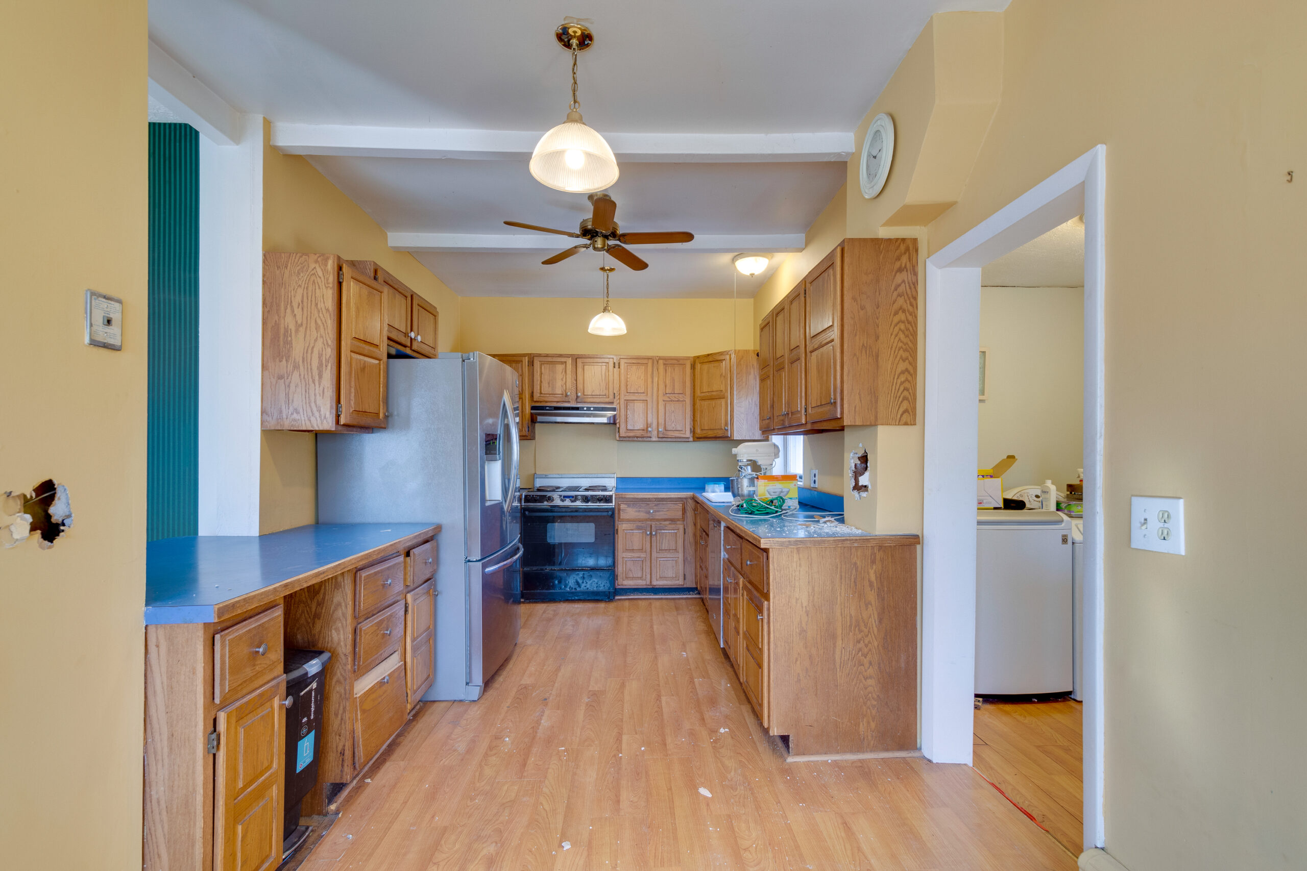 Image of a kitchen in a vintage New England home before renovation, featuring old wooden cabinets, blue countertops, and outdated appliances. The yellow walls and worn flooring further highlight the need for a comprehensive update to bring the space up to modern standards.