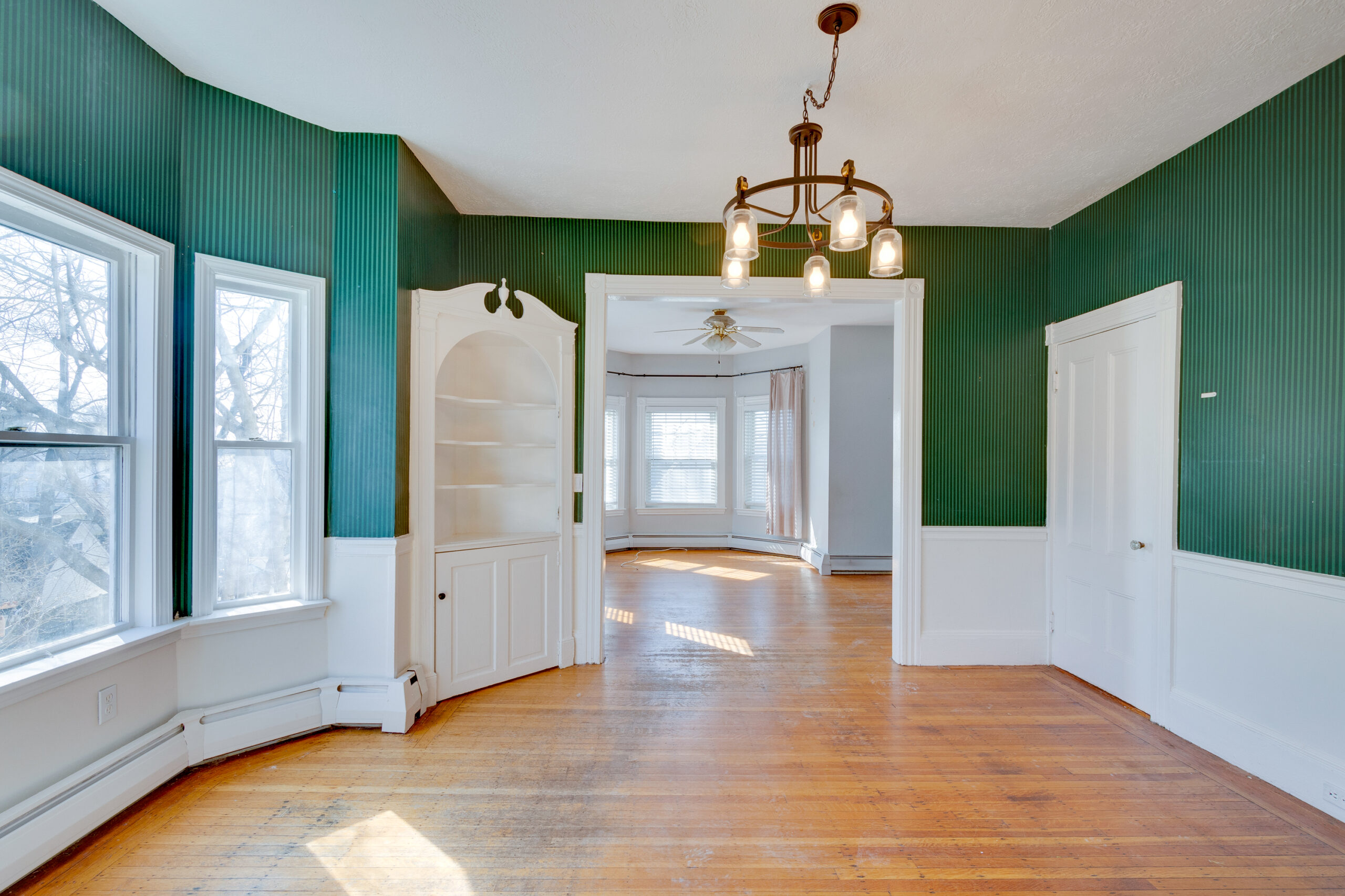 Image of a bay window area in the dining room of a vintage New England home before renovation. The space features dark green wallpaper above white wainscoting and a built-in corner cabinet. The bay windows allow ample natural light to flood the room, which has worn hardwood floors and an old chandelier. The adjacent room with yellow walls indicates a dated and eclectic interior style, emphasizing the need for renovation.