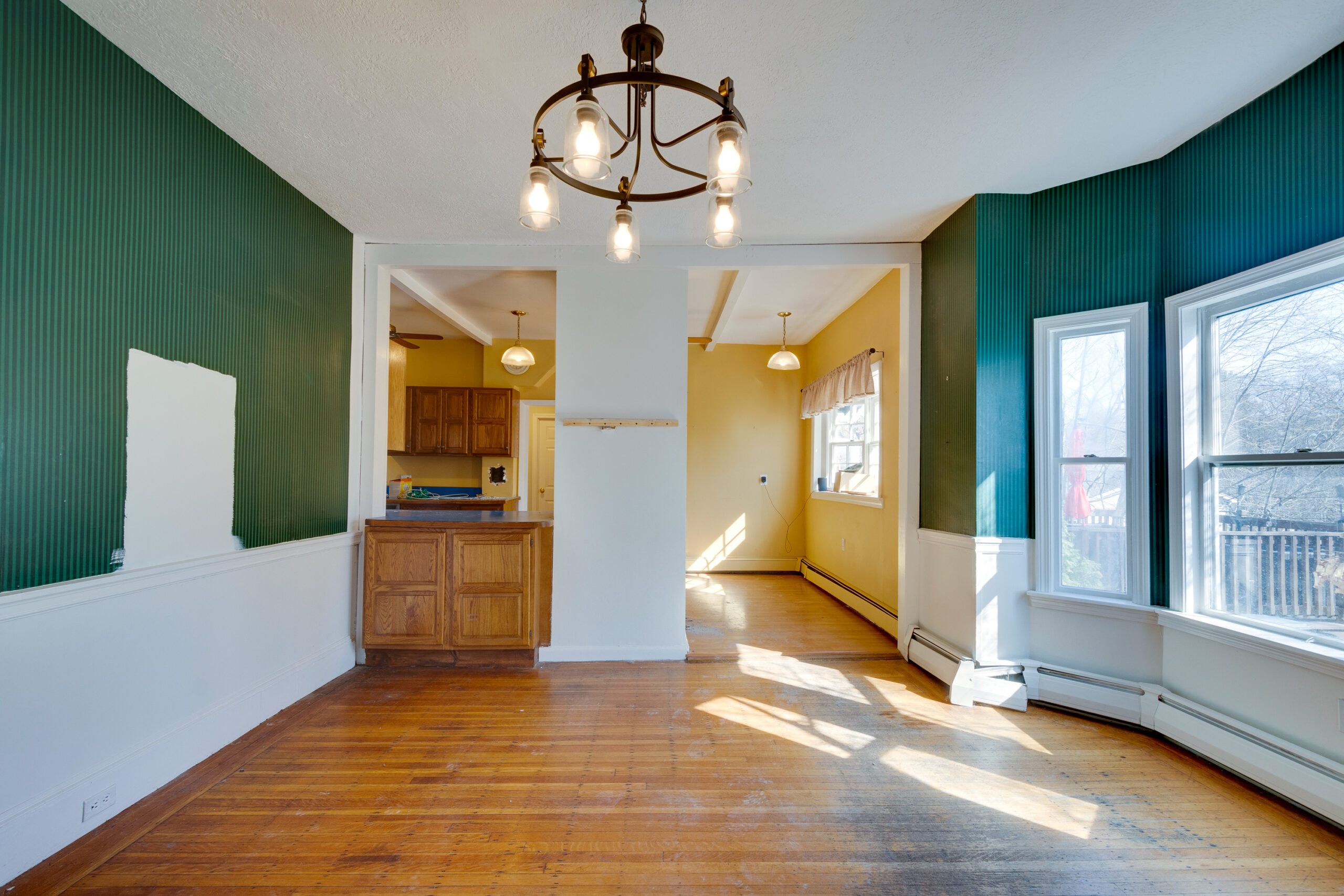 Image of a dining room before renovation in a vintage New England home. The room features outdated green and yellow walls, a dark wood chandelier, and large windows letting in natural light. The hardwood floors are scuffed, and there is a visible patch on the green wall, indicating the beginning of the renovation process.