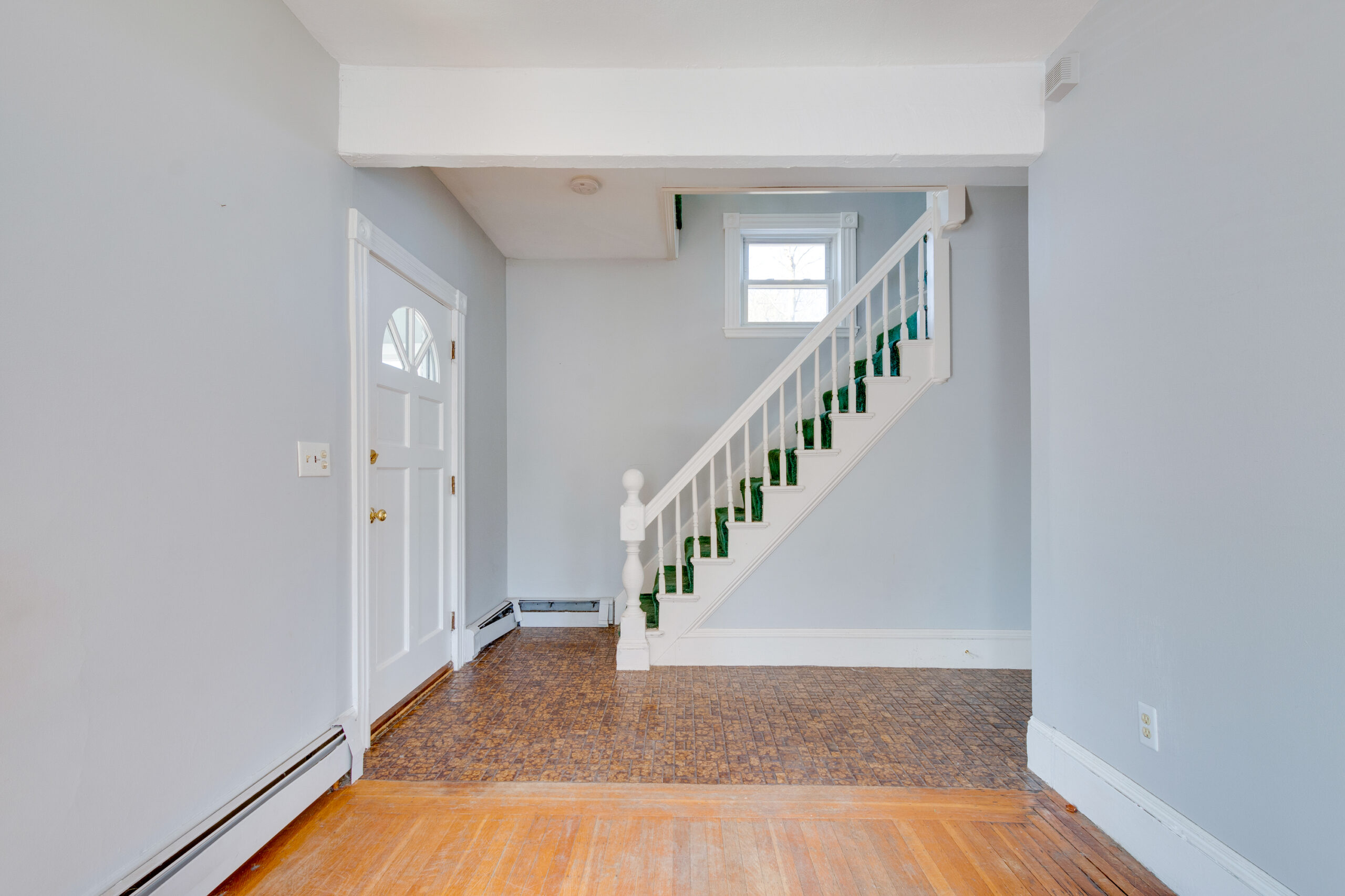 Image of a dated foyer featuring mismatched flooring, dull grey walls, and dark green carpeted stairs. The lighting is inadequate, making the space feel cramped and uninviting. Historic home over 100 years old in New England.