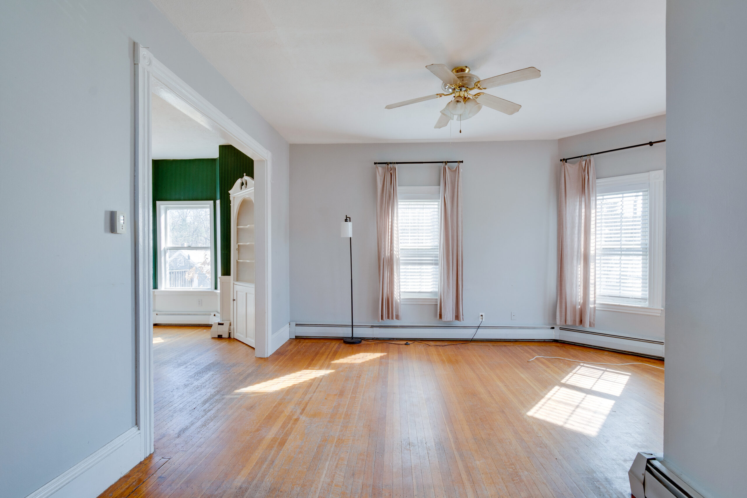 Image of a vintage New England living room before renovation. The room features large windows with pink curtains, outdated light grey walls, and worn hardwood floors. A ceiling fan with outdated lighting and a floor lamp contribute to the room's dated appearance. The space, with its traditional wood trim and baseboard heating, reflects the home's historical charm.