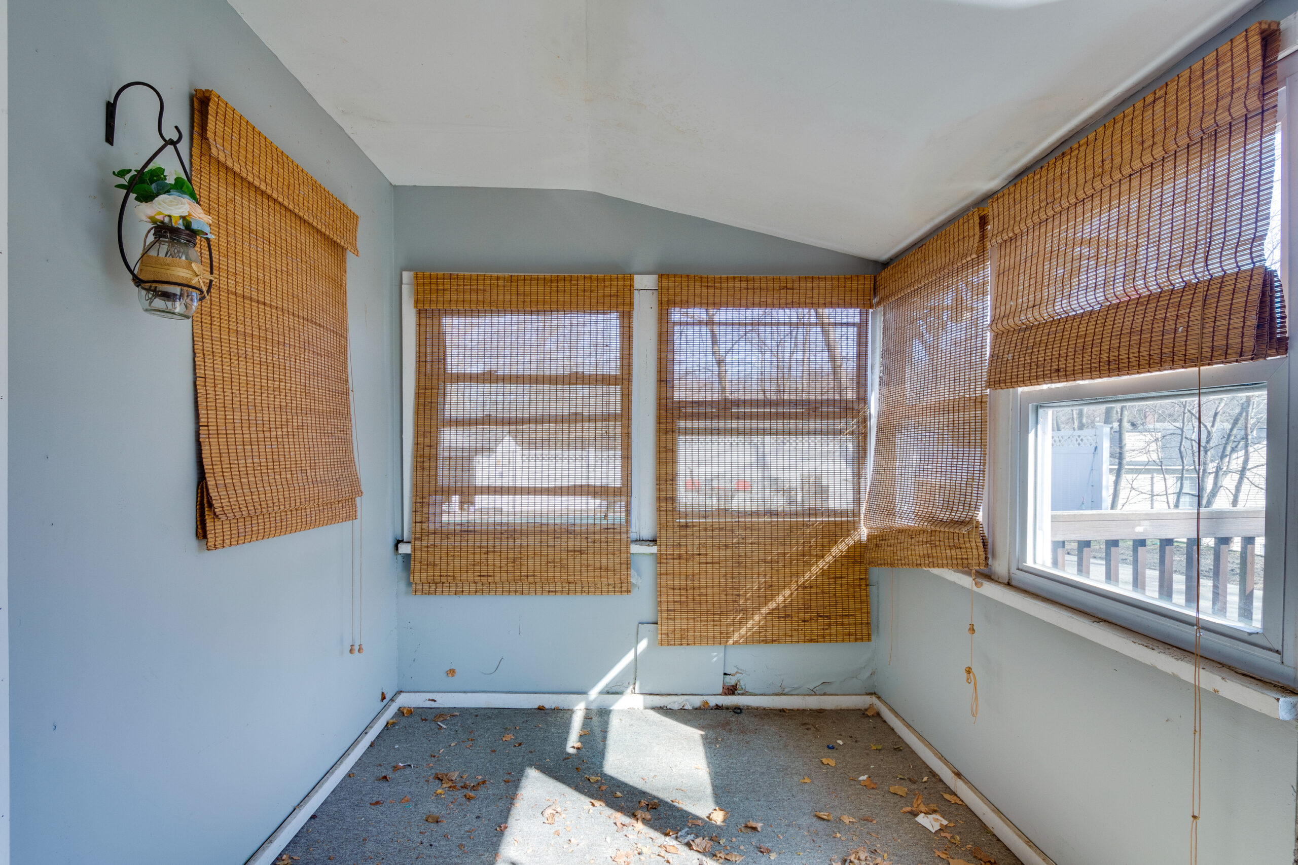 Image of a sunroom before renovation in a vintage New England home. The room has worn-out bamboo blinds partially covering large windows, a light blue wall, and a slightly stained carpet. A decorative lantern hangs on the wall, and the room appears to be in need of a thorough makeover.