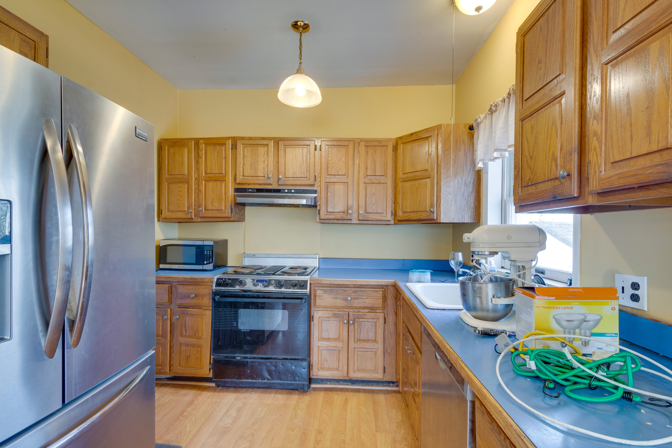 mage of a kitchen in a vintage New England home before renovation. This view shows a large hole in the yellow wall, old wooden cabinets, and blue countertops. The room also has a ceiling fan, outdated appliances, and a mix of old flooring, indicating significant wear and tear.