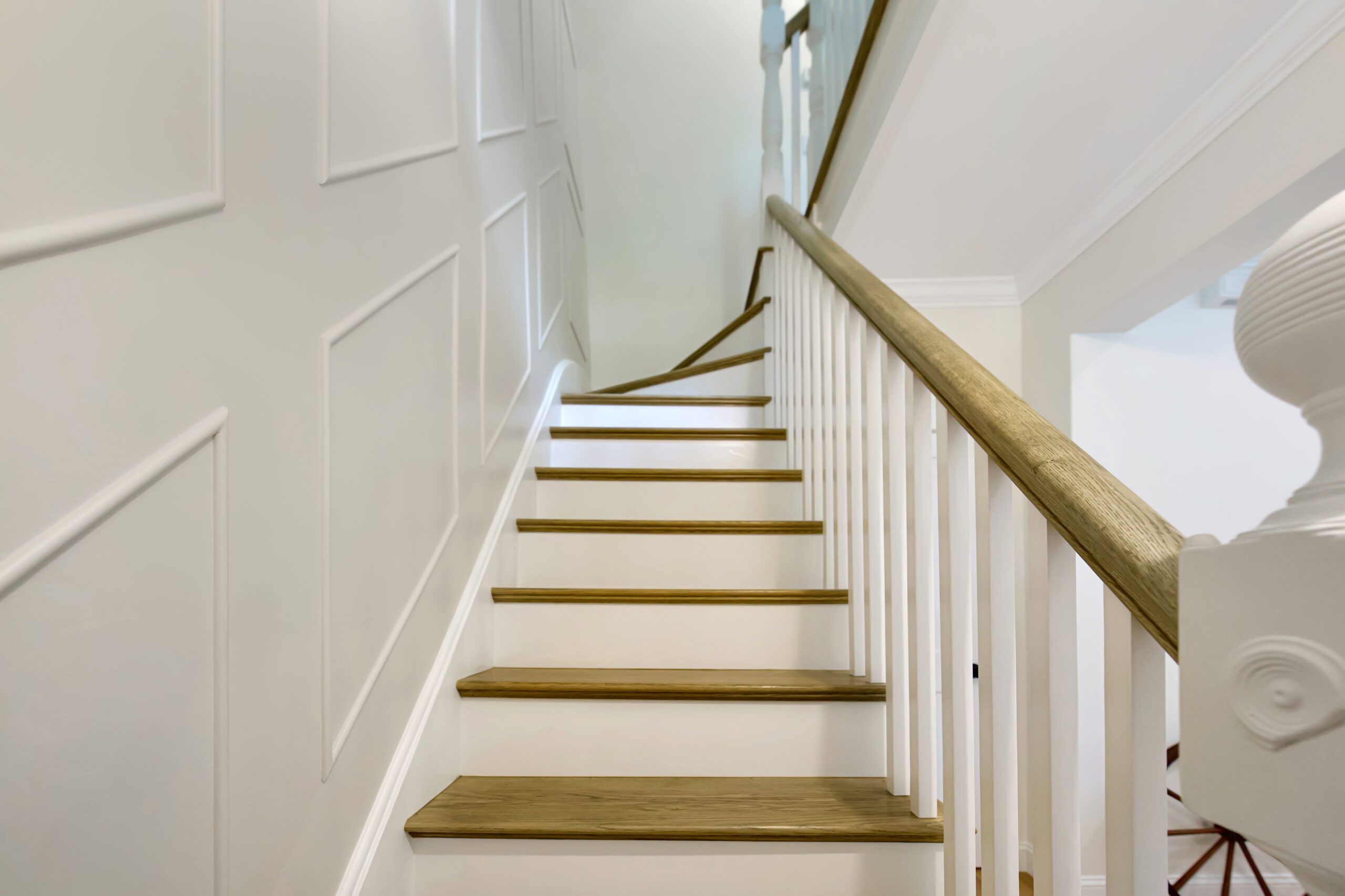 Image of a renovated staircase with polished wooden steps and white risers. The crisp white railing complements the new wall paneling, adding texture and interest to the space, illuminated by an elegant light fixture. Original post was restored.