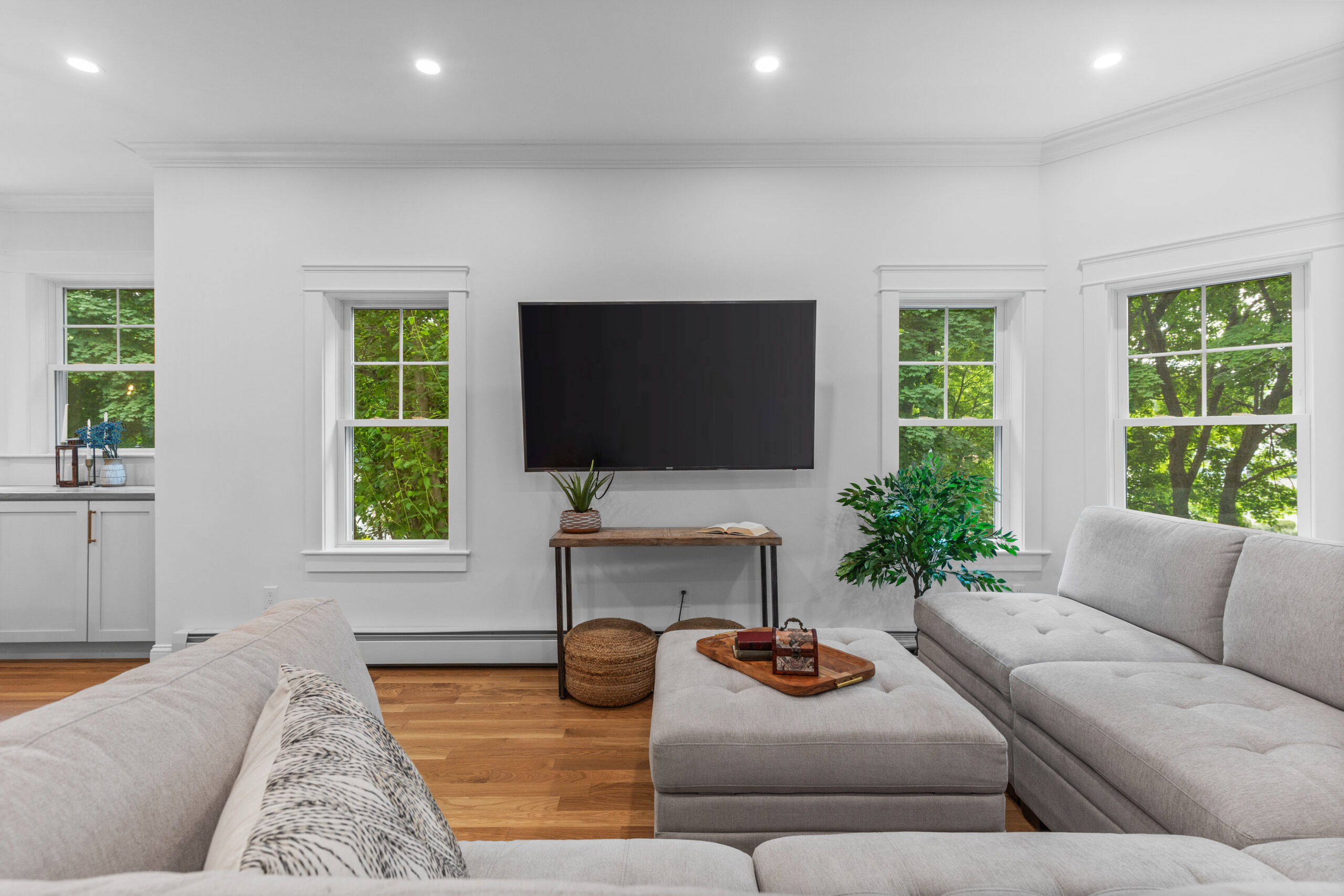 Image of a newly renovated living room in a vintage New England home. The room features a wall-mounted flat-screen TV between two large windows, allowing natural light to illuminate the space. A cozy sectional sofa with neutral-colored upholstery and decorative pillows faces the TV. The hardwood flooring adds warmth, while a side table with potted plants and woven baskets underneath enhances the modern yet comfortable aesthetic. The walls are painted white, providing a clean and bright atmosphere.
