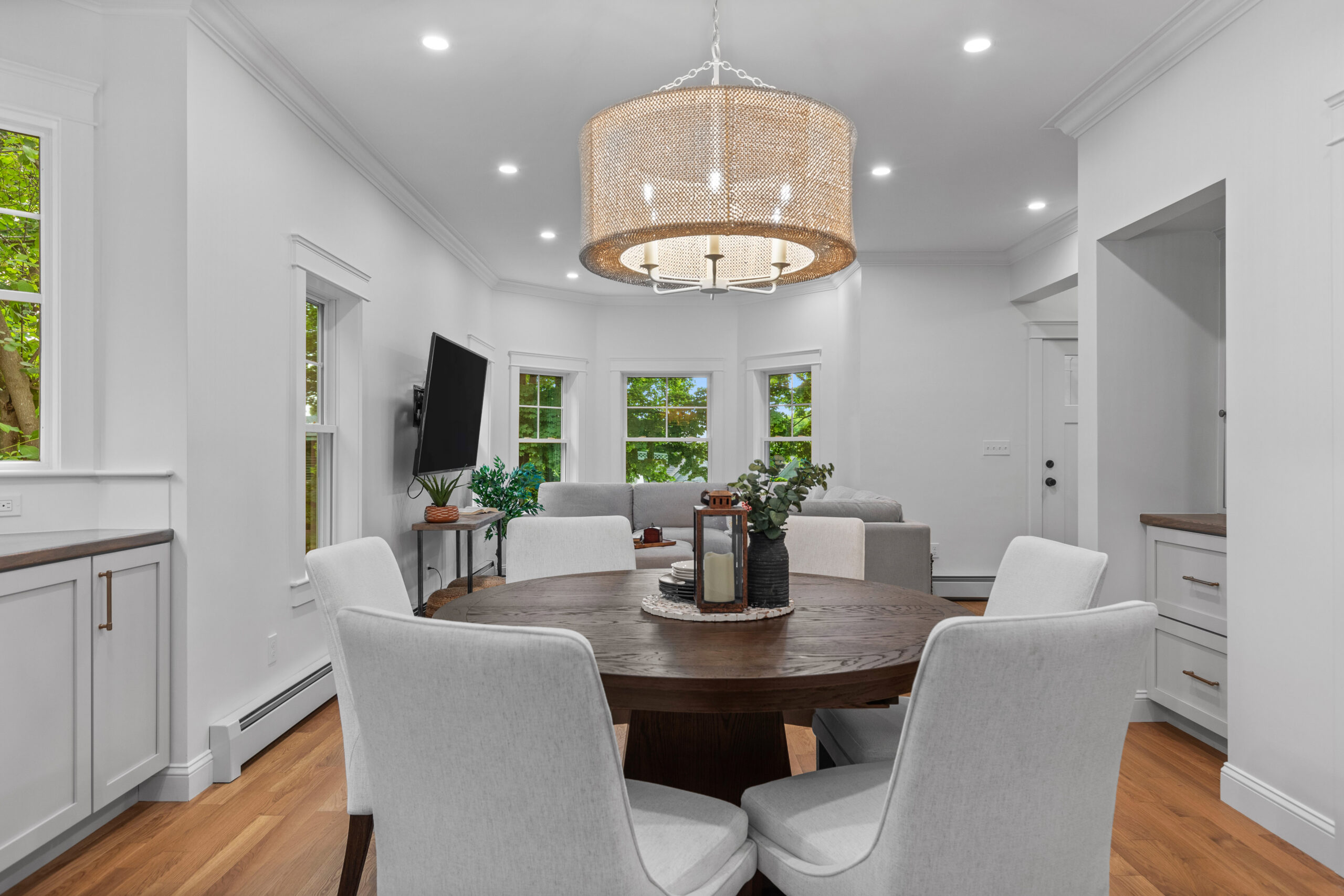 Image of a beautifully renovated living and dining room in a vintage New England home. The space features custom cabinetry with sleek wooden countertops and modern hardware. White walls and large windows allow natural light to brighten the room, showcasing the new hardwood flooring and contemporary decor, creating a harmonious blend of classic and modern design.