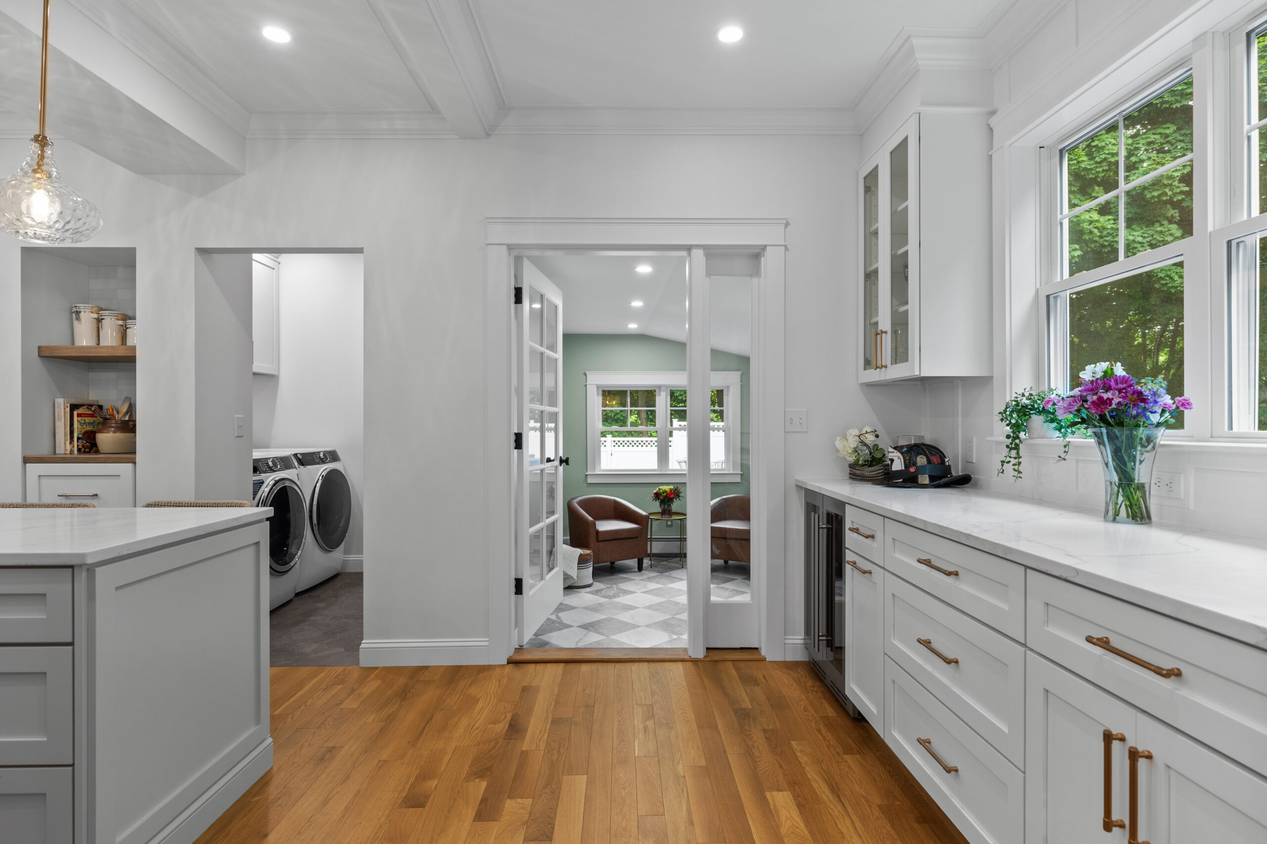 Image of a beautifully renovated kitchen in a vintage New England home, showcasing the open view into a sunroom. The kitchen features white cabinetry, marble countertops, and hardwood floors. French doors lead to the sunroom, which has been updated with green walls and modern furniture, creating a seamless and inviting transition between spaces.