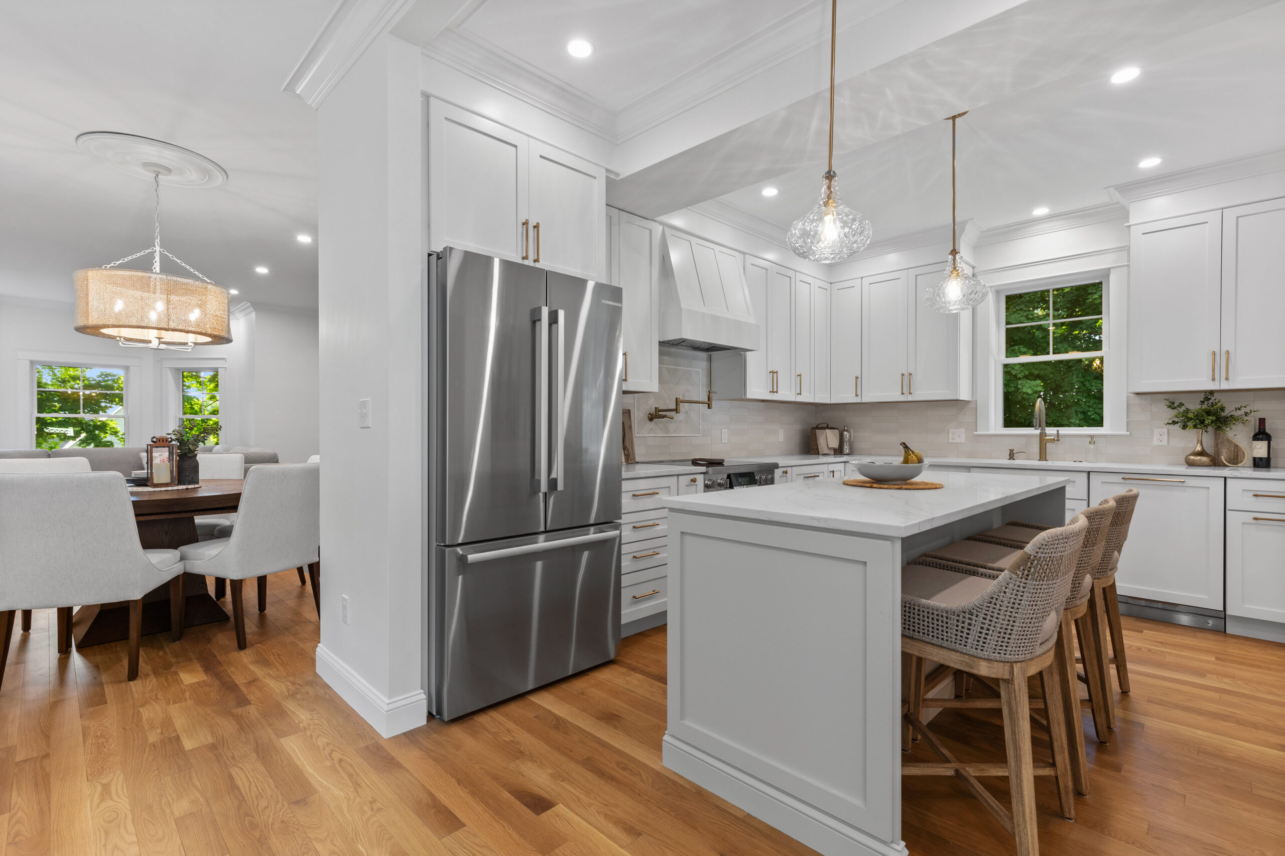 Image of a modern kitchen in a vintage New England home, post-renovation. The kitchen boasts an expansive layout with white cabinetry, marble countertops, and a central island. Elegant pendant lights and under-cabinet lighting highlight the clean lines and contemporary design, while hardwood floors add warmth to the space.