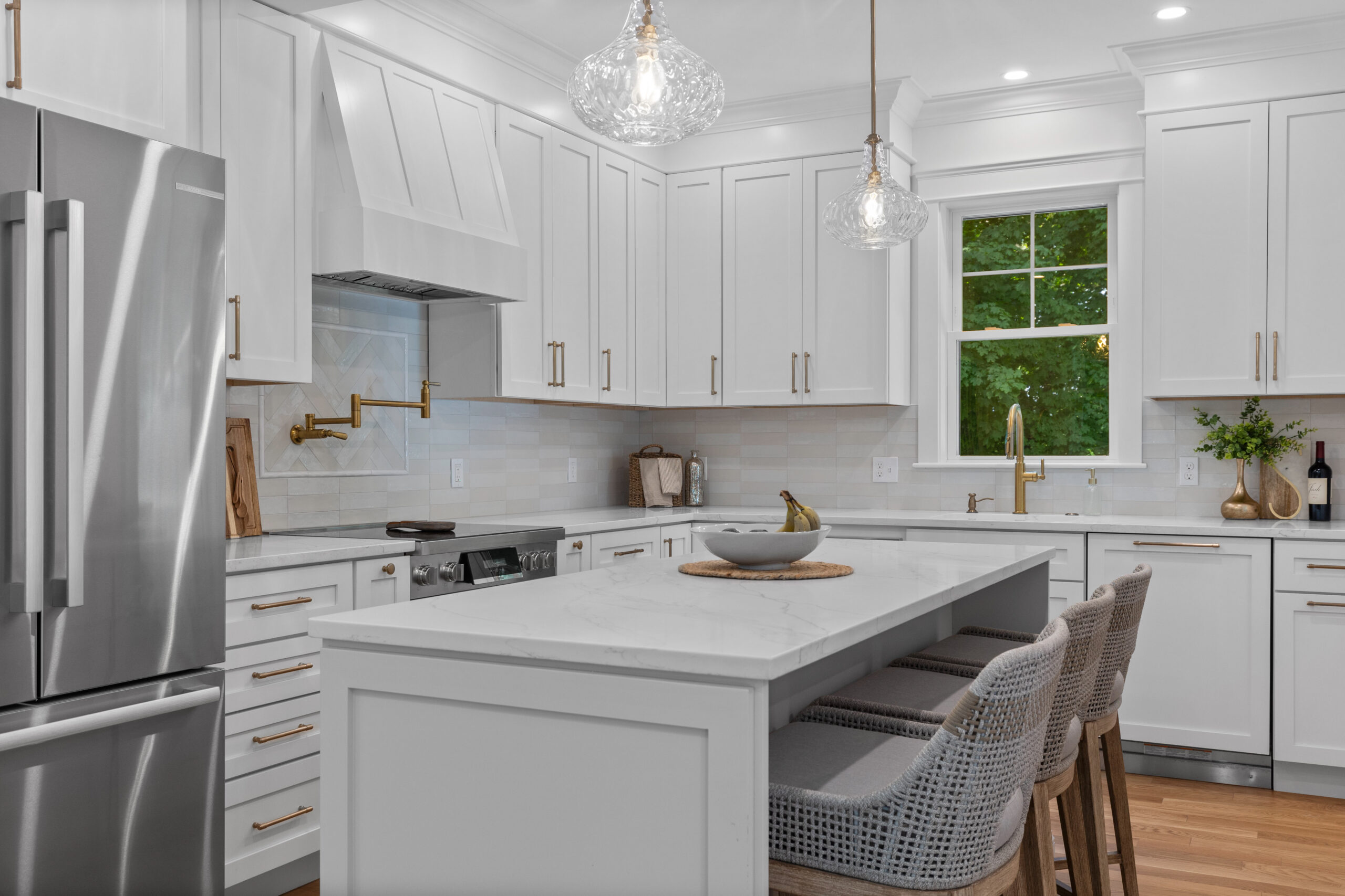 Image of a beautifully renovated kitchen in a vintage New England home. The kitchen features a large island with seating, white shaker cabinets, and stainless steel appliances. The space is illuminated by pendant lights and recessed lighting, with natural light streaming in from large windows, creating a bright and welcoming atmosphere.