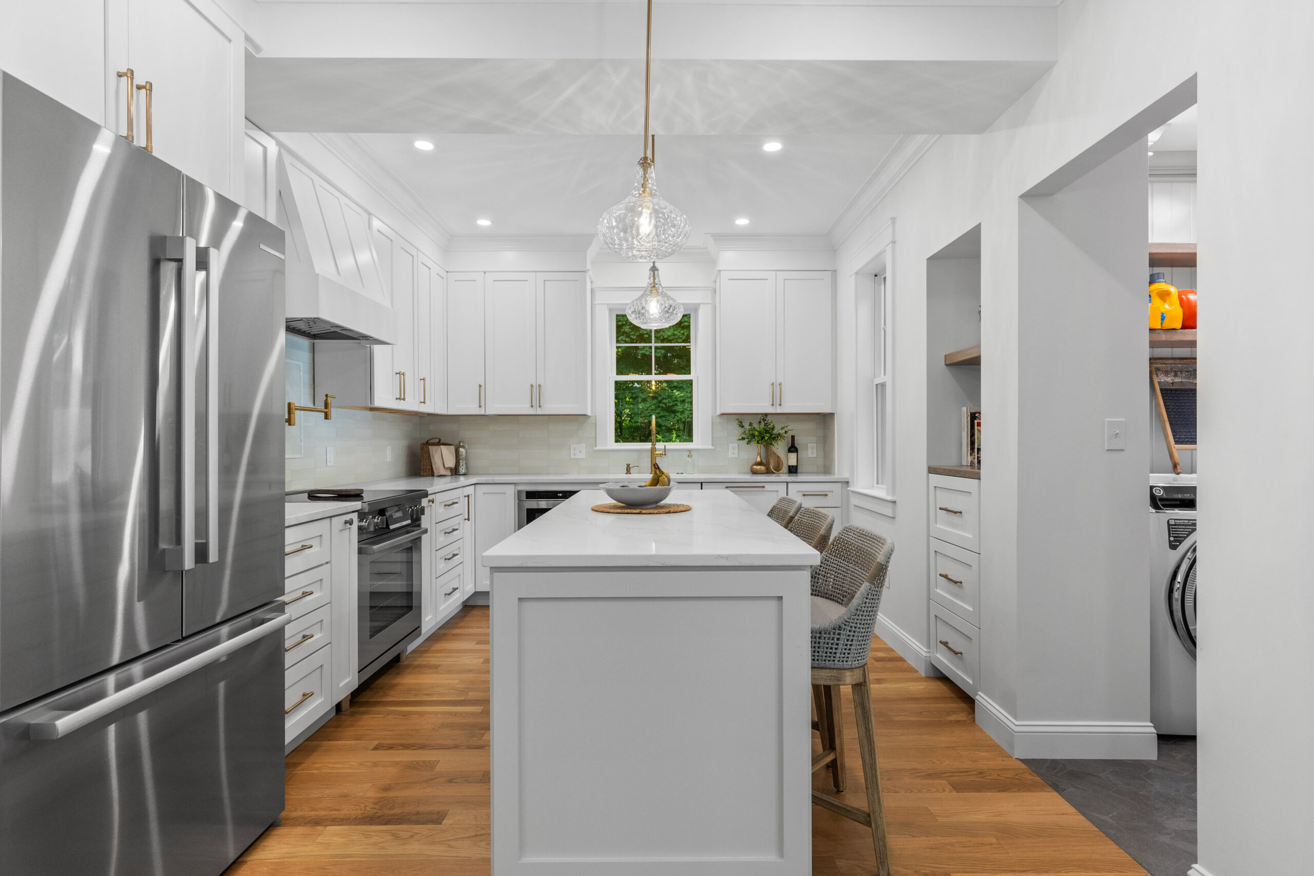 Image of a modern kitchen in a vintage New England home, post-renovation. The kitchen boasts an expansive layout with white cabinetry, marble countertops, and a central island. Elegant pendant lights and under-cabinet lighting highlight the clean lines and contemporary design, while hardwood floors add warmth to the space.