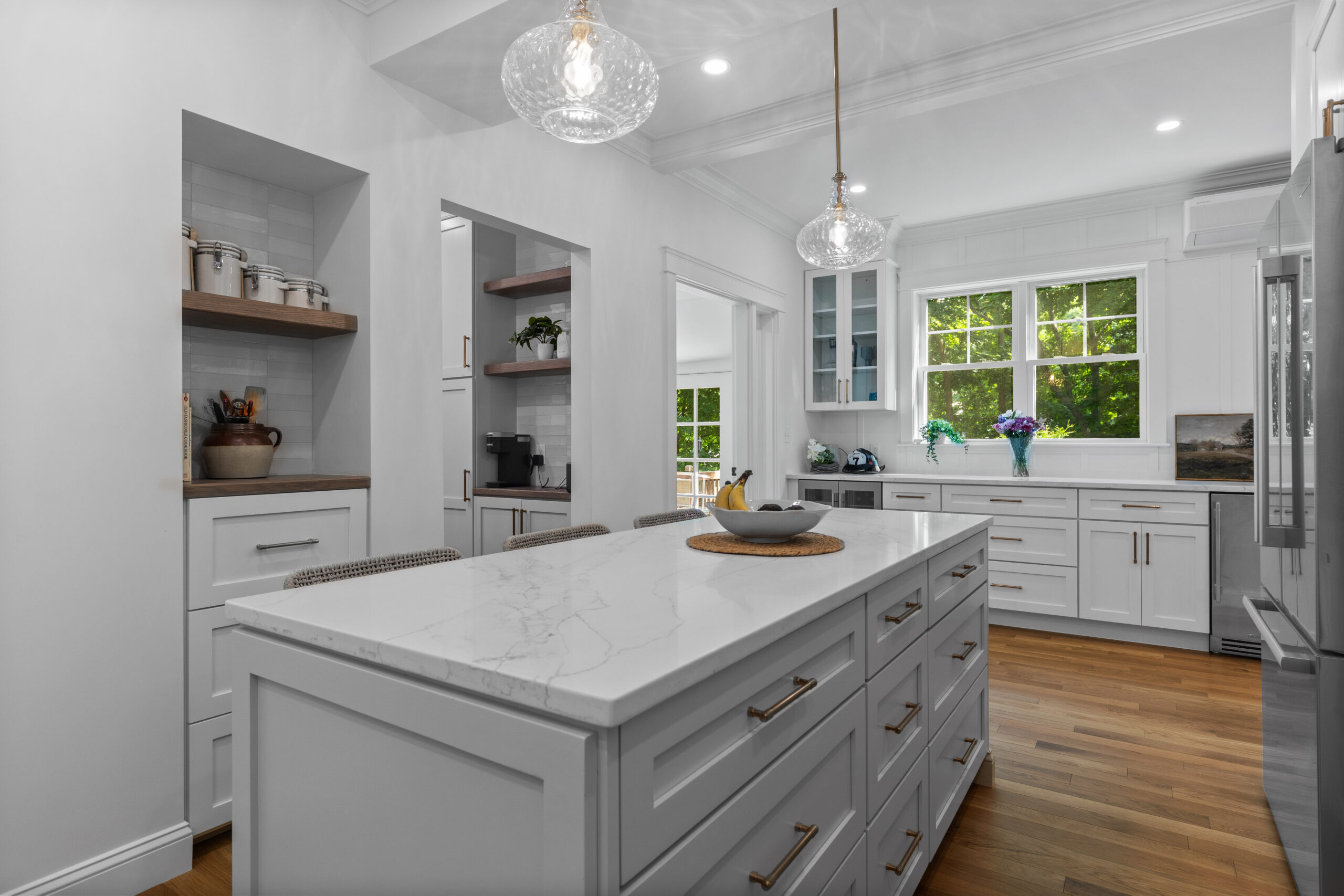 Image of a newly renovated kitchen in a vintage New England home, highlighting the long countertop with white cabinetry and brass hardware. The bright and airy kitchen opens up to a sunroom, which has comfortable seating and large windows that bring in plenty of natural light. Fresh flowers and decorative items add a homely touch to the modern space