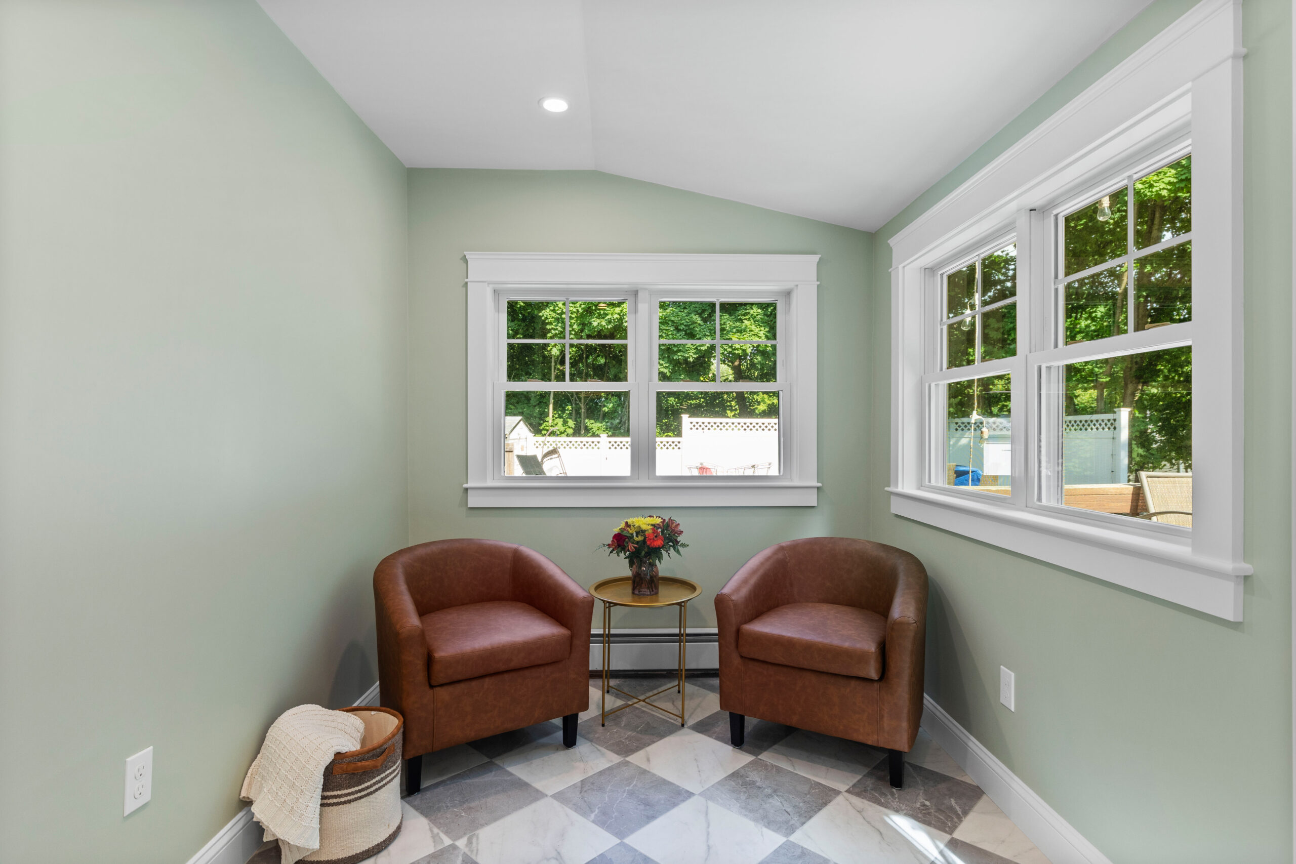 Image of a fully renovated sunroom in a vintage New England home. The sunroom features light green walls, large windows, and marble tile flooring. It is furnished with two brown leather armchairs and a small round table with a floral arrangement, providing a cozy and bright space to relax and enjoy the view of the backyard.