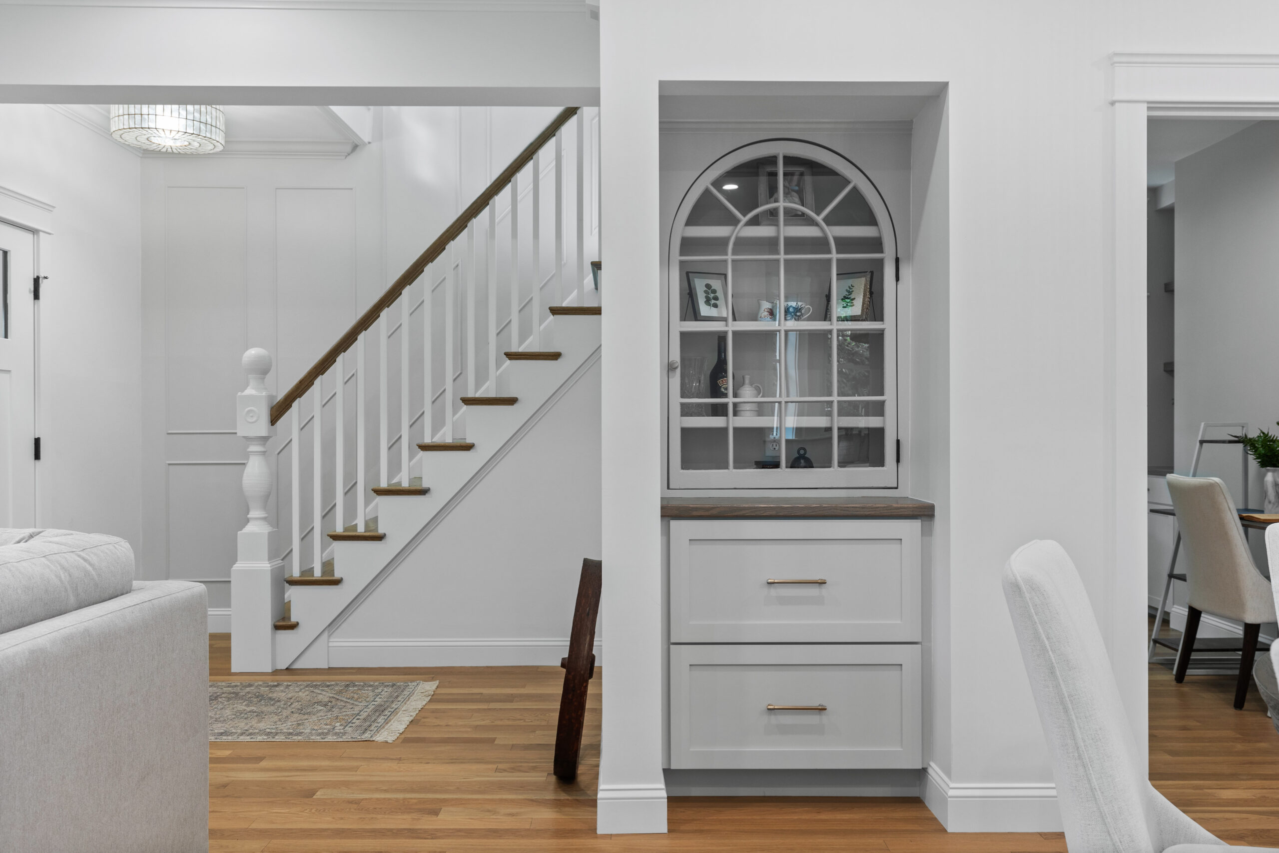 Image of an elegantly renovated space in a vintage New England home, featuring a built-in display cabinet with glass doors and wooden shelves. The entryway showcases updated hardwood flooring, white walls, and modern fixtures, including a chic light fixture and a decorative rug. The adjacent office area is visible through a pair of French doors, completing the sophisticated and functional design.