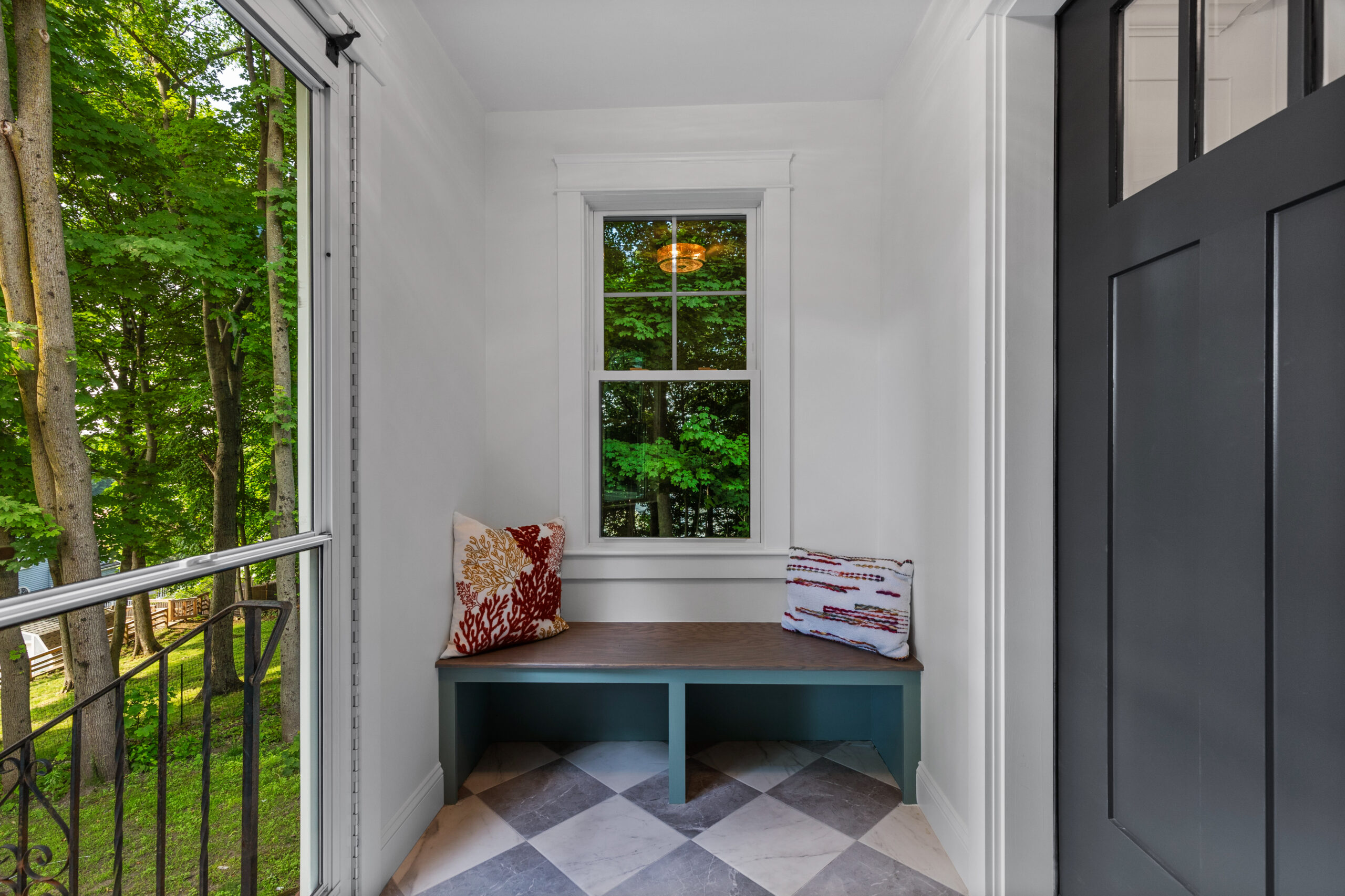 Image of a charming mudroom bench painted in Sherwin Williams Jade Dragon, complemented by a checkerboard marble floor. The bench, adorned with decorative pillows, offers a cozy seating area beneath a large window that provides a scenic view of lush greenery outside. The black door - Benjamin Moore graphite- adds a striking contrast, enhancing the overall aesthetic of this inviting and practical space.
