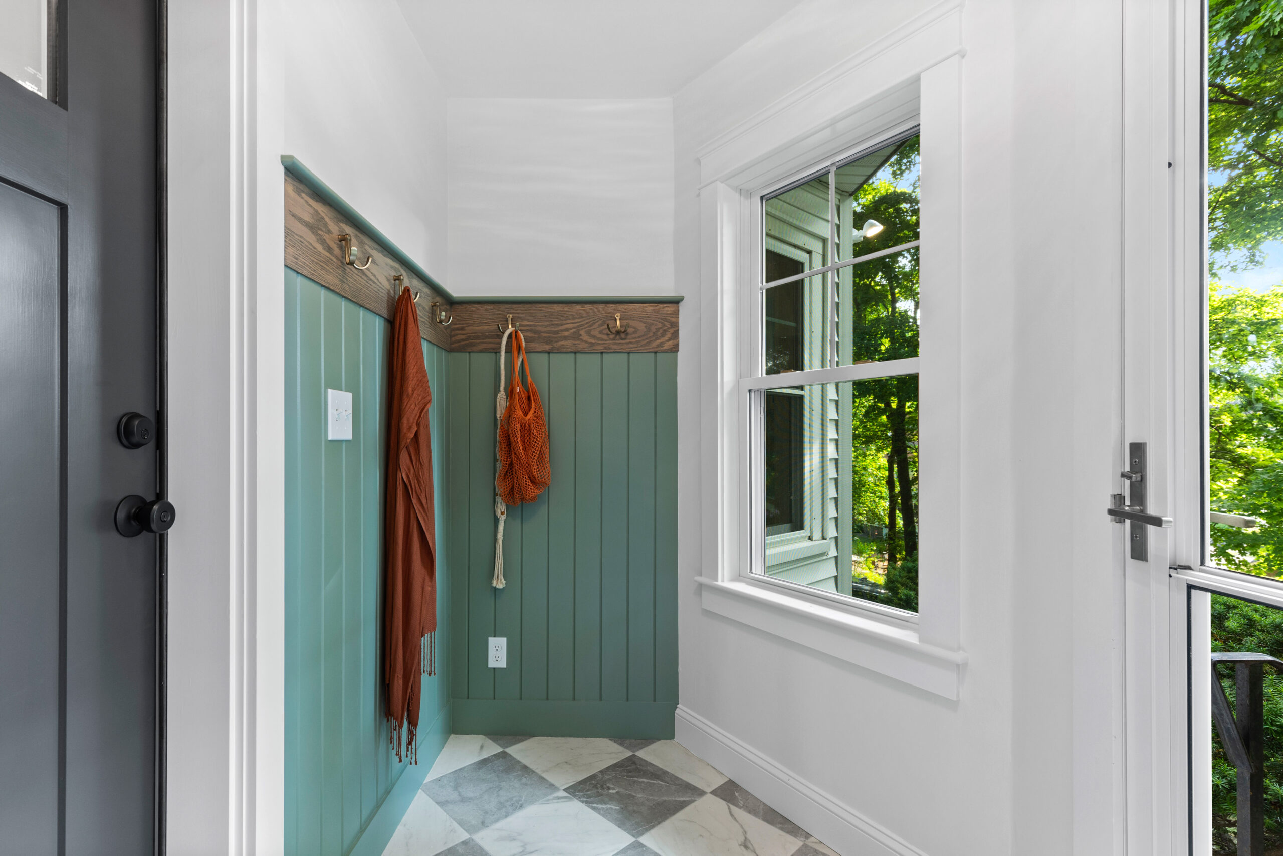 Image of a newly renovated mudroom featuring Sherwin Williams Jade Dragon paint on the wainscoting. The space includes elegant wooden hooks for hanging items, with an accent of natural wood trim. The checkerboard marble floor adds a touch of luxury, and a large window floods the room with natural light, highlighting the stylish and functional design.