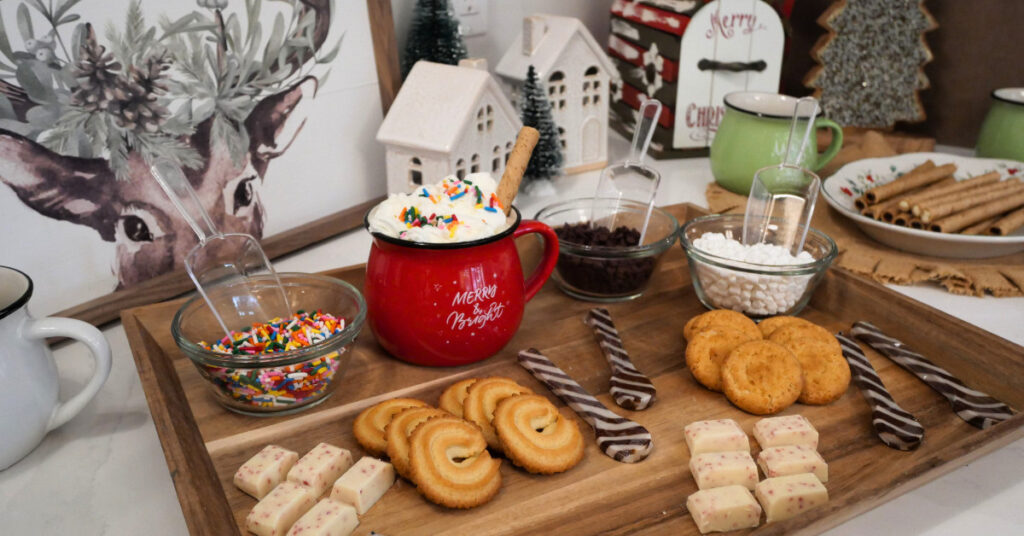 A holiday hot chocolate bar setup featuring a red cocoa mug with whipped cream and sprinkles surrounded by bowls of toppings, cookies, chocolate peppermint sticks, and festive Christmas décor including mini houses and trees.