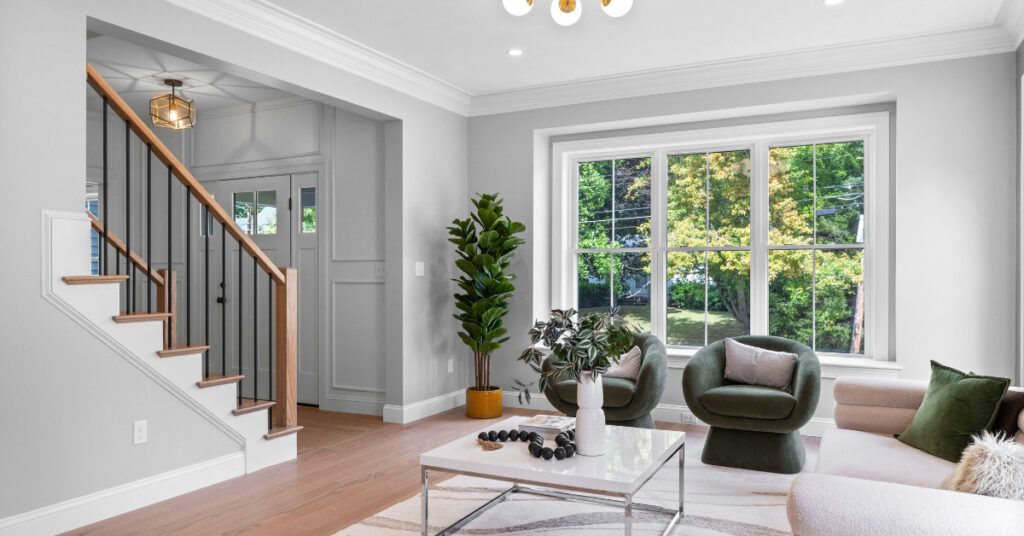 A bright, modern living room designed by Kadilak Homes in Burlington, Massachusetts, featuring Benjamin Moore Collingwood paint on the walls, large windows letting in natural light, green accent chairs, and a sleek staircase with black metal balusters.