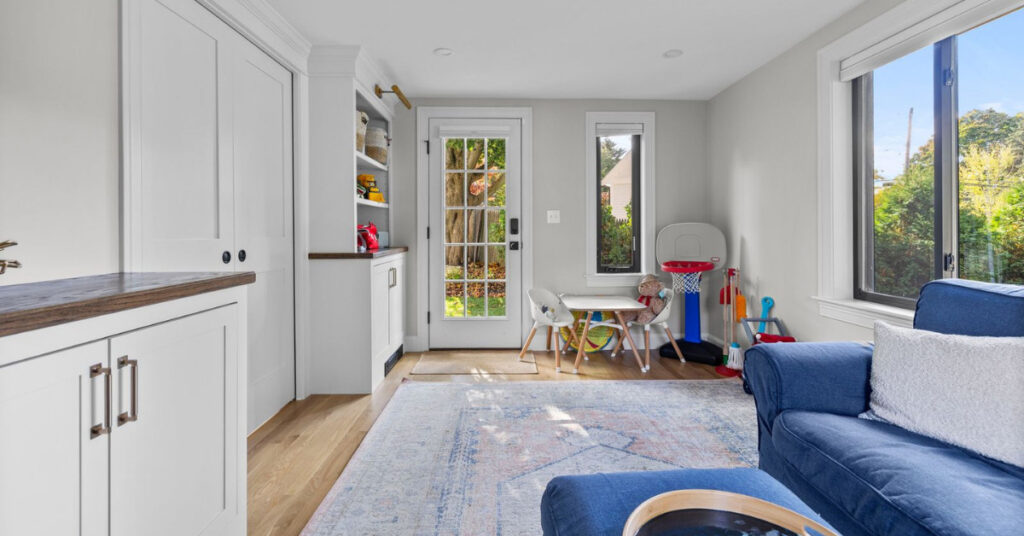 Bright playroom with built-in white cabinets, wood countertops, and large windows allowing natural light. A kids' play table and basketball hoop are set against a cozy, functional design.