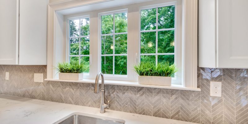 This kitchen portrays a contemporary design featuring a large window that bathes the room in natural light, offering a view of lush greenery outside. The window trim and framing are painted white, matching the walls and creating a fresh and clean aesthetic. Below the window, the backsplash is adorned with grey chevron tiles, contributing a touch of elegance and texture to the space. On the windowsill, there are two rectangular planters with vibrant green plants, adding a pop of color and life to the scene. The countertop, boasting a marble-like finish, extends seamlessly to a deep, under-mount sink with a modern stainless-steel faucet, indicating functionality paired with style. The presence of electrical outlets on the backsplash suggests convenience for kitchen appliances, while the absence of visible clutter indicates a minimalist and tidy space, likely designed to appeal to those who appreciate a blend of modern design and natural elements in their living environment.