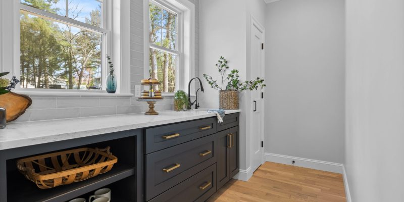 "A bright, elegant walk in pantry space with navy blue cabinets and a full sink and large windows offering a view of trees and a clear blue sky. Below the windows is a white marble countertop with a dark navy cabinetry, adorned with brass handles. An assortment of decorative items, including a wooden cake stand with a glass dome, a rustic wooden bowl, and a basket with greenery, adds a homely touch. The kitchen features a farmhouse-style sink with a modern black faucet, and neatly arranged on an open shelf below the countertop are matching white mugs in a row. The room is completed with light wood flooring and a simple yet stylish black wall sconce