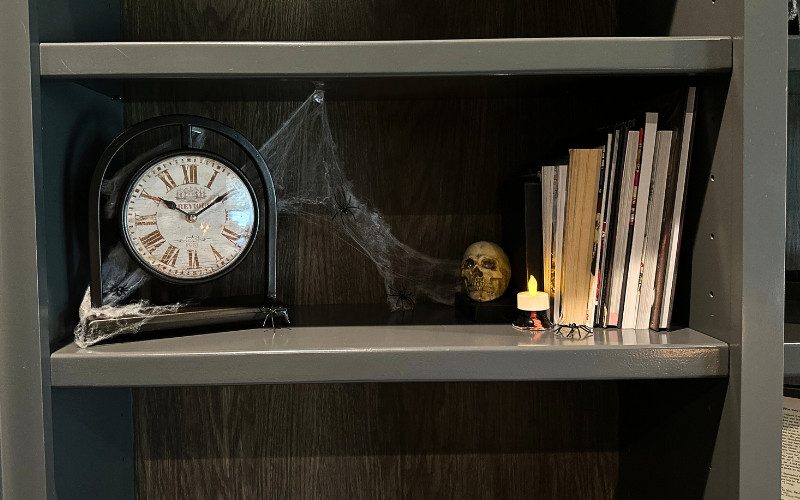 Dark wooden shelves displaying a vintage clock, faux cobwebs, a small skull, and thrifted books in a haunted library setting.