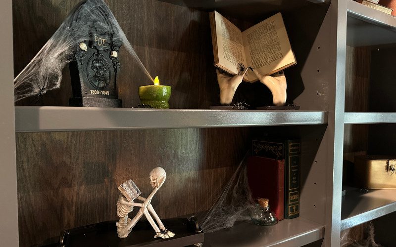 Dark wooden shelves with a tombstone honoring Edgar Allan Poe, flanked by faux cobwebs and a glowing candle in a dark academia-style home library.