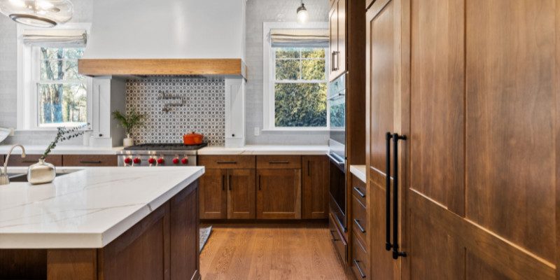 A modern farmhouse-style kitchen featuring rich wooden cabinetry and a large central island with a white quartz countertop. The kitchen's focal point is a striking patterned tile backsplash behind the stove, framed by a white range hood with a wooden trim. The space is illuminated by natural light streaming through two windows, complemented by stylish pendant lighting above the island. The design balances warmth and elegance, combining rustic wood tones with contemporary finishes for a welcoming and functional cooking space.