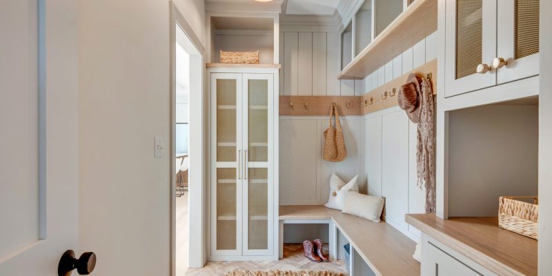 A tidy and welcoming mudroom featuring a wooden bench with beige cushions, nestled between white storage cabinets. Above the bench, a shelf with brass hooks holds a woven bag and a scarf, while a wicker basket sits atop the cabinet, adding a touch of rustic charm. A pair of boots are neatly placed under the bench. The walls are painted in a soft, light grey, and the herringbone-patterned floor adds a classic touch. A warm pendant light hangs from the ceiling, illuminating the space with a cozy glow. Through the open glass-paneled door on the left, a glimpse into another room with similar interior colors can be seen, providing a sense of continuity throughout the home. Benjamin Moore cumulous cloud 1550