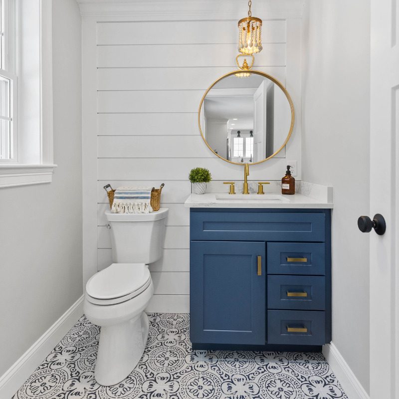 Coastal bathroom design with white horizontal shiplap wall paneling, statement light pendant, circular gold vanity mirror, Sherwin Williams Indigo Batik bathroom vanity cabinets, and a statement tile in a black and white damask pattern.