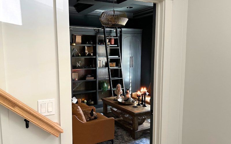 A view into a cozy haunted library with burnt orange leather chairs, flickering candles, and dark wood shelves filled with vintage and spooky decor.