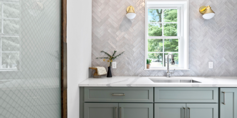 Light & airy walk-in pantry design with light sage green kitchen cabinetry, textured subway tile in a chevron pattern, white quartz countertops, gold hardware and fixtures, and a large window to let in all the natural light.