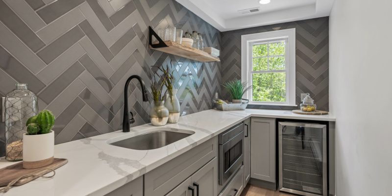 Walk in pantry with glossy and gray herringbone tile backsplash. Walk in pantry with oak flooring, wine refrigerator, and sink.