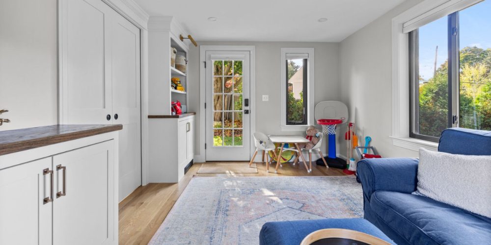 Bright playroom with built-in white cabinets, wood countertops, and large windows allowing natural light. A kids' play table and basketball hoop are set against a cozy, functional design.
