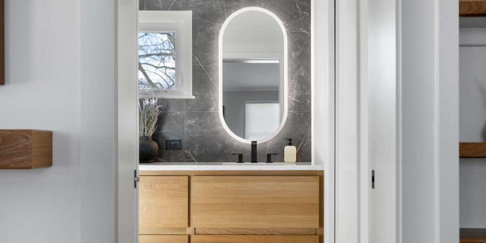 Modern bathroom vanity with natural wood cabinetry, illuminated oval mirror, black fixtures, and under-cabinet toe-kick lighting, viewed from a hallway entrance.