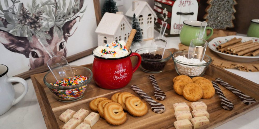 A holiday hot chocolate bar setup featuring a red cocoa mug with whipped cream and sprinkles surrounded by bowls of toppings, cookies, chocolate peppermint sticks, and festive Christmas décor including mini houses and trees.