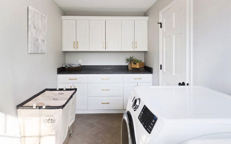 White laundry room with built-in countertops and overhead cabinets for organized folding space by Kadilak Homes.