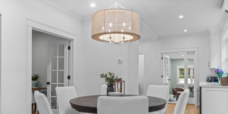 This image showcases a cozy dining area with a round, dark wood dining table surrounded by white upholstered chairs. The room features a large, drum-shaped chandelier with a textured beige shade, which is perfectly proportioned to the size of the table and the space. The chandelier adds a warm, inviting glow to the room, complementing the bright white walls and creating a balanced, intimate atmosphere. The clean, simple lines of the room’s design, including the white trim and glass-paneled doors leading to adjacent rooms, enhance the modern yet comfortable aesthetic. This image illustrates the importance of choosing a lighting fixture that fits both the size and style of a dining space.