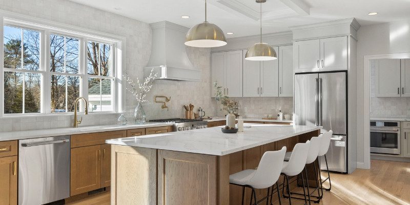 This image depicts a bright and modern kitchen with a large center island featuring a marble countertop and seating for four with white barstools. Above the island, two oversized, dome-shaped pendant lights in a brushed gold finish provide focused illumination, perfectly complementing the sleek and airy design of the space. The kitchen is fitted with white upper cabinets, light wood lower cabinets, and stainless steel appliances, creating a clean, cohesive look. Large windows above the sink flood the space with natural light, enhancing the warm, welcoming atmosphere. This image highlights the importance of selecting appropriately scaled lighting fixtures to enhance both the functionality and aesthetic of a spacious kitchen