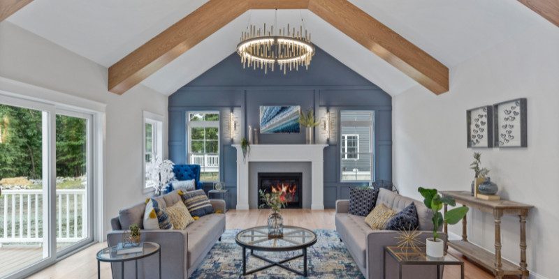 spacious and elegant living room with a vaulted ceiling featuring exposed wooden beams. The focal point is a modern, circular chandelier suspended from the ceiling, perfectly proportioned to the room's scale. The walls are painted in soft, neutral tones, with a feature wall in a rich blue hue behind a white fireplace. The room is furnished with two gray sofas facing each other, accented with blue and yellow throw pillows, and a glass-topped coffee table in the center. Large windows on one side of the room allow natural light to flood the space, creating a bright and inviting atmosphere. This image demonstrates how selecting the right size lighting fixture can enhance the balance and aesthetic of a large living area